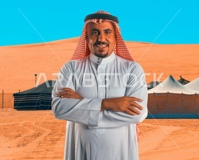 صورة لرجل عربي خليجي سعودي، ينظر إلى الكاميرا بأيدي متقاطعة مع خلفية لخيمة في صحراء المملكة العربية السعودية، الكثبان الرملية، رحلة برية في المناطق الصحراوية بالسعودية، السياحة في السعودية، الطبيعة الصحراوية في السعودية، صحاري السعودية