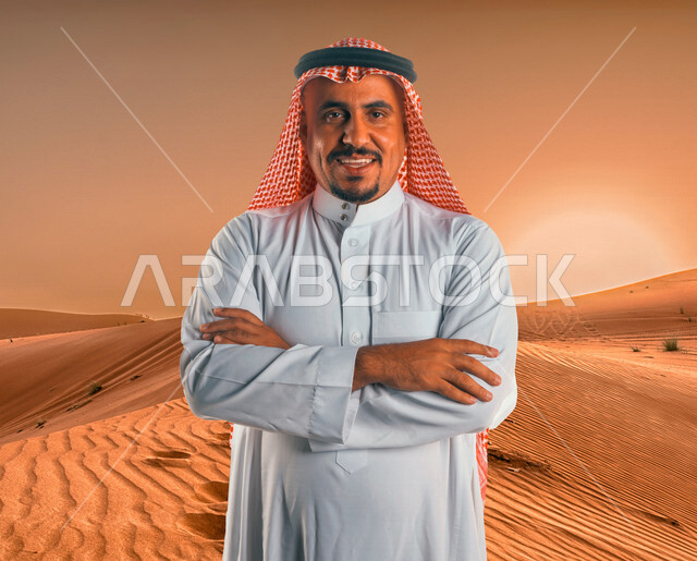 صورة لرجل عربي خليجي سعودي، ينظر إلى الكاميرا بأيدي متقاطعة مع خلفية لصحراء المملكة العربية السعودية، الكثبان الرملية، رحلة برية في المناطق الصحراوية بالسعودية، السياحة في السعودية، الطبيعة الصحراوية في السعودية، صحاري السعودية