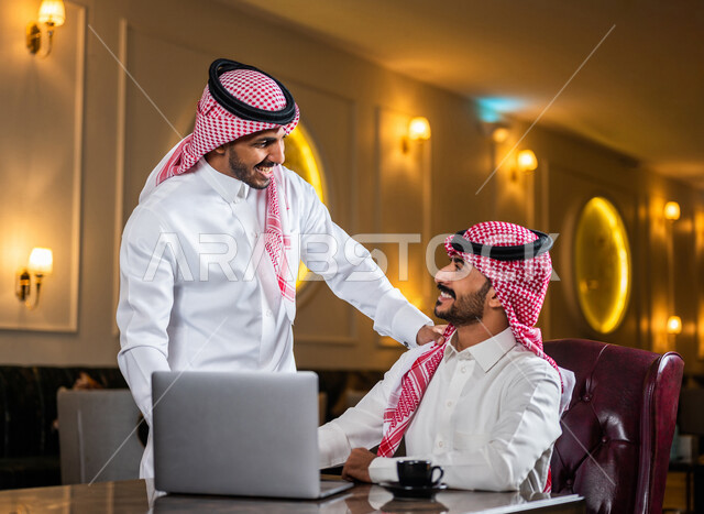 زملاء عمل عرب سعوديون خليجيون مجتمعون في المقهى, حوار ونقاش عمل خارج الشركه, انجهاز مهام العمل عبر جهاز الحاسوب, إدارة الأعمال عن بعد, توزيع مهام العمل بين الزملاء,  الإستمتاع بشرب القهوه في متجر القهوه, مقاهي في المملكه العربيه السعوديه