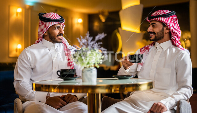 رجلان عربيان خليجيان سعوديان مبتسمان يجلسان في المقهى يقومان بشرب فنجان القهوة الساخنة ، جلسة صداقة ، لقاء و تعارف ، حوار و نقاش ، علاقات إجتماعية ، قضاء أوقات ممتعة