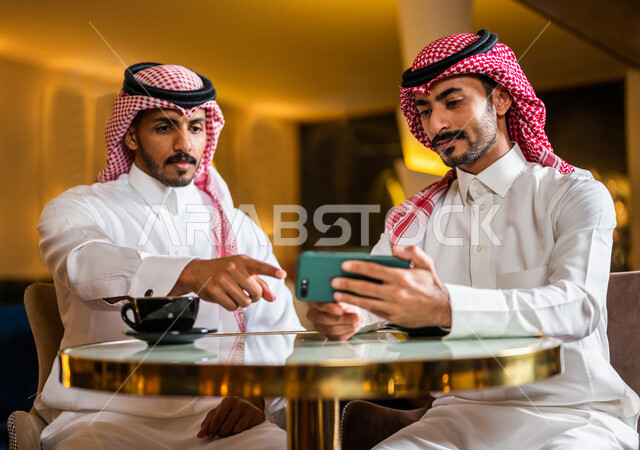 رجلان عربيان خليجيان سعوديان مبتسمان يجلسان في المقهى ، يستمتعان بشرب القهوة الساخنة و مشاهدة البرامج الترفيهية على الهاتف المحمول ، رجل سعودي يشير بيده تجاه الهاتف ، ديوانية شباب السعودية ، جلسة صداقة ، أجواء شبابية في مقهى سعودي