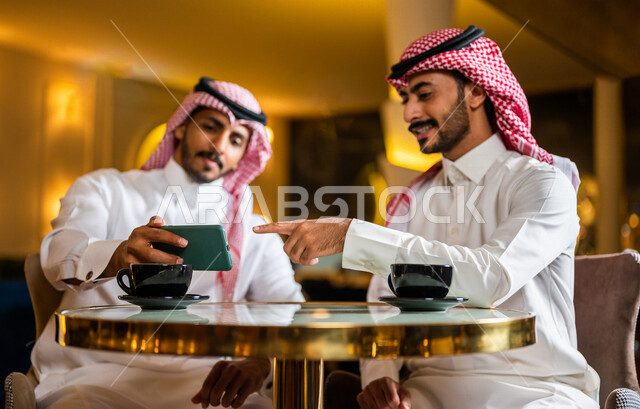 رجلان عربيان خليجيان سعوديان مبتسمان يجلسان في المقهى ، يستمتعان بشرب القهوة الساخنة و مشاهدة البرامج الترفيهية على الهاتف المحمول ، رجل سعودي يشير بيده تجاه الهاتف ، ديوانية شباب السعودية ، جلسة صداقة ، أجواء شبابية في مقهى سعودي
