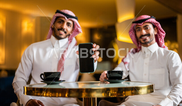 رجلان عربيان خليجيان سعوديان مبتسمان يجلسان في المقهى ، رجل سعودي يقوم بعرض الهاتف المحمول بسعادة ، عرض الخدمات التطبيقية ، تطبيق المطاعم ، قائمة المنيو الالكترونية عبر تطبيقات الهاتف المحمول ، التسويق و الاعلان