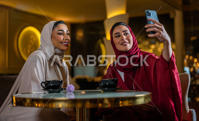 سيدتان عربيتان خليجيتان سعوديتان مبتسمتان تجلسان في المقهى  تقومان بإستخدام الهاتف المحمول و شرب فنجان القهوة الساخنة ، مشاهدة برنامج ترفيهي ، أخذ صورة تذكارية ، إجراء مكالمة فيديو ، جلسة صداقة ، علاقات إجتماعية ، قضاء أوقات ممتعة 