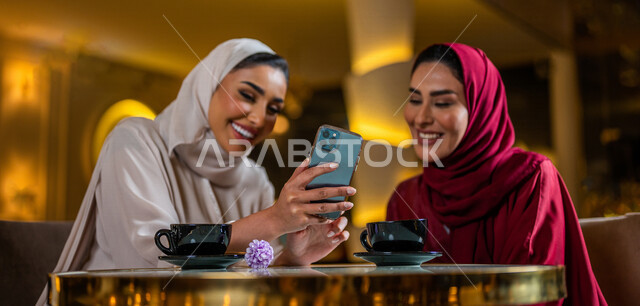 Two smiling Saudi Arabian Gulf women sitting in a café using a mobile phone and drinking a cup of hot coffee, watching an entertainment program, taking a souvenir photo, making a video call, friendship session, social relations, spending fun times