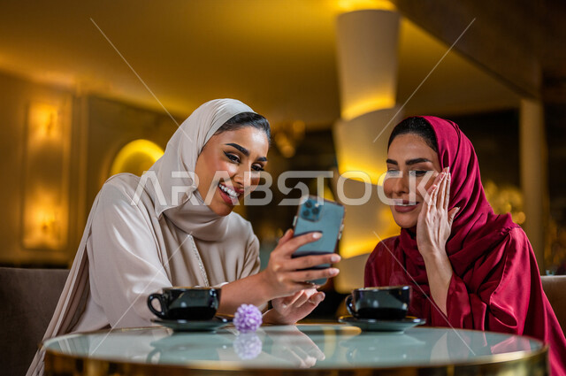 سيدتان عربيتان خليجيتان سعوديتان مبتسمتان تجلسان في المقهى  تقومان بإستخدام الهاتف المحمول و شرب فنجان القهوة الساخنة ، مشاهدة برنامج ترفيهي ، أخذ صورة تذكارية ، إجراء مكالمة فيديو ، جلسة صداقة ، علاقات إجتماعية ، قضاء أوقات ممتعة 