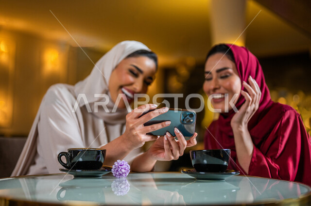 سيدتان عربيتان خليجيتان سعوديتان مبتسمتان تجلسان في المقهى  تقومان بإستخدام الهاتف المحمول و شرب فنجان القهوة الساخنة ، مشاهدة برنامج ترفيهي ، أخذ صورة تذكارية ، إجراء مكالمة فيديو ، جلسة صداقة ، علاقات إجتماعية ، قضاء أوقات ممتعة 