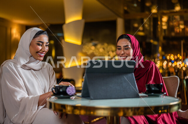 سيدتان عربيتان خليجيتان سعوديتان مبتسمتان تجلسان في المقهى ، تقومان بحجز الطلبات من خلال التابلت ، استخدام تطبيق المطعم لعرض المنيو ، قائمة طعام رقمية ، جلسة صداقة ، علاقات إجتماعية ، قضاء أوقات ممتعة 