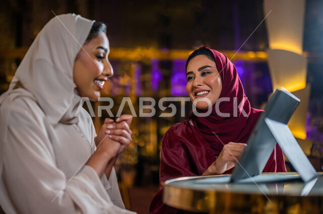 سيدتان عربيتان خليجيتان سعوديتان مبتسمتان تجلسان في المقهى ، تقومان بحجز الطلبات من خلال التابلت ، استخدام تطبيق المطعم لعرض المنيو ، قائمة طعام رقمية ، جلسة صداقة ، علاقات إجتماعية ، قضاء أوقات ممتعة 