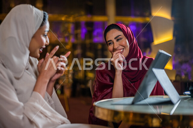 Two smiling Saudi Arabian Gulf women sitting in the café, booking orders through the tablet, using the restaurant application to display the menu, a digital menu, a friendship session, social relations, spending fun times