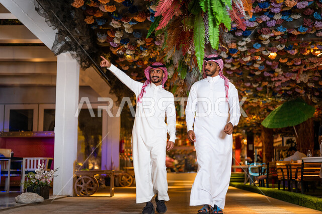 رجلان عربيان خليجيان سعوديان صديقان مبتسمان يسيران معاً في نزهة ممتعة لإحدى الأماكن الترفيهية ، رجل سعودي  يشير باتجاه مكان ما بسعادة ، صداقات و تعارف ، علاقات إجتماعية ، قضاء أوقات ممتعة