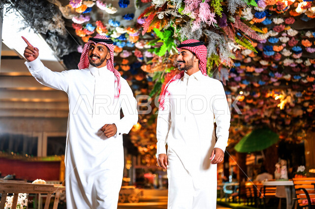 رجلان عربيان خليجيان سعوديان صديقان مبتسمان يسيران معاً في نزهة ممتعة لإحدى الأماكن الترفيهية ، رجل سعودي  يشير باتجاه مكان ما بسعادة ، صداقات و تعارف ، علاقات إجتماعية ، قضاء أوقات ممتعة