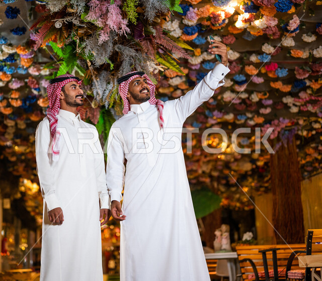 رجلان عربيان خليجيان سعوديان صديقان مبتسمان يقومان بإلتقاط صورة سلفي تذكارية عبر الهاتف المحمول ،  صداقات و تعارف ، علاقات إجتماعية ، قضاء أوقات ممتعة