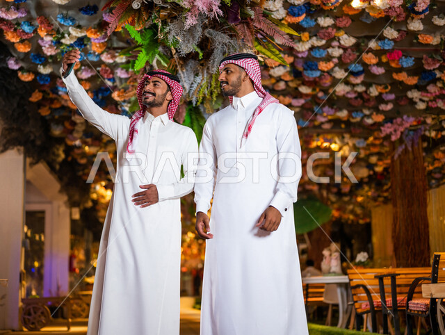 رجلان عربيان خليجيان سعوديان صديقان مبتسمان يقومان بإلتقاط صورة سلفي تذكارية عبر الهاتف المحمول ،  صداقات و تعارف ، علاقات إجتماعية ، قضاء أوقات ممتعة