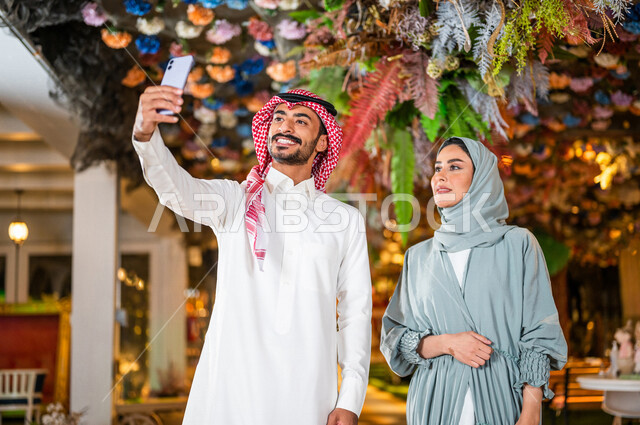 زوجان عربيان خليجيان سعوديان مبتسمان يلتقطان صورة سيلفي تذكارية بالهاتف المحمول ، يسيران معاً في نزهة ممتعة لإحدى الأماكن الترفيهية ، التنزه في وقت العطلة ، المشاركة بفعاليات ممتعة ، قضاء أوقات عائلية ممتعة 
