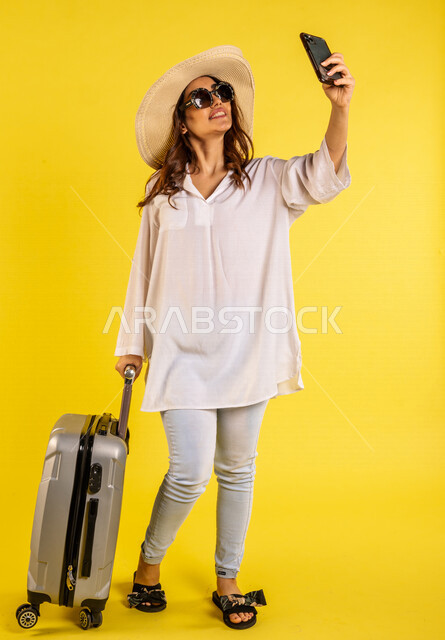 Portrait of a Saudi Arabian Gulf woman wearing casual clothes for summer trips with her travel bag next to her, taking memorial selfies through a mobile phone, enjoying filming the tourist trip, video call, full-length picture, full body
