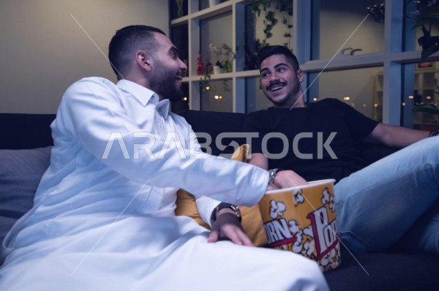 Two Saudi Arabian Gulf youth enjoy watching a movie on the TV, a group home entertainment activity, facial gestures indicating integration and interaction with the movie, spending a good time with friends, enjoying eating popcorn