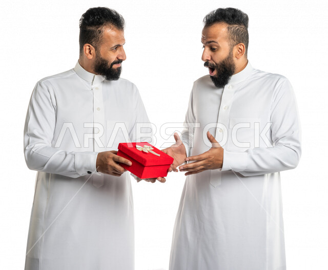 بورتريه لرجل عربي خليجي سعودي يقدم صندوق أحمر لأخاه التوأم بايماءات تدل على الدهشة والسعادة ، هدية عيد ميلاد ، هدايا تذكارية ، المشاركة في الأعياد والإحتفالات ، مناسبات سعيدة