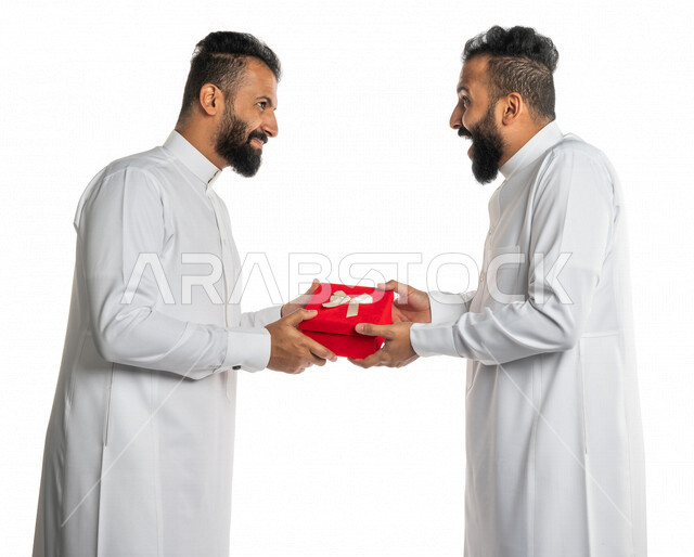 بورتريه لرجل عربي خليجي سعودي يقدم صندوق أحمر لأخاه التوأم بايماءات تدل على الدهشة والسعادة ، هدية عيد ميلاد ، هدايا تذكارية ، المشاركة في الأعياد والإحتفالات ، مناسبات سعيدة