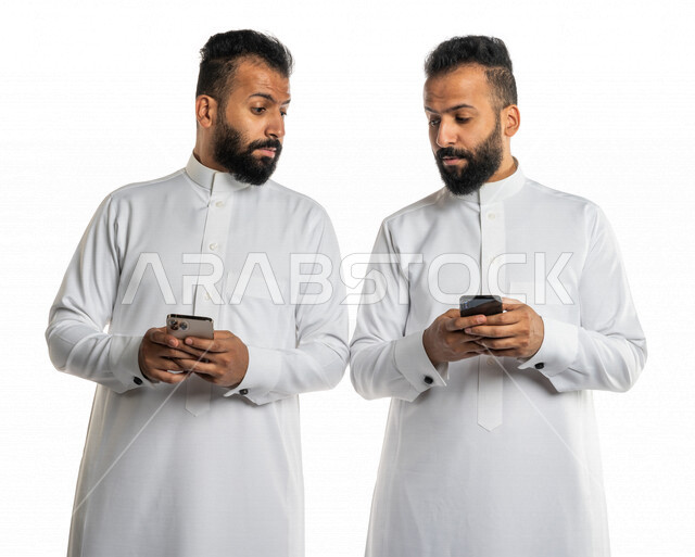 Portrait of two Saudi Arabian Gulf twin men using a mobile phone, peeking at the other phone, surveillance and intrusion, eavesdropping on personal phones, internet addiction, white background