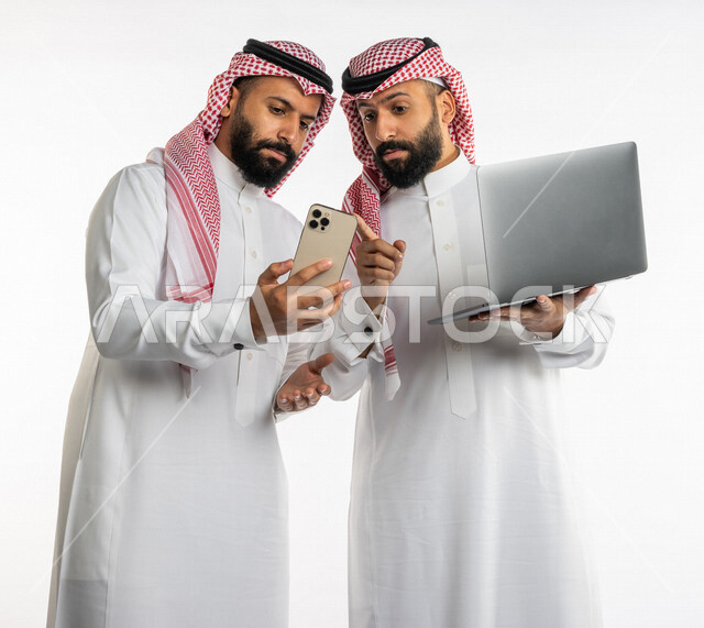 بورتريه لرجلان توأم عربيان خليجيان سعوديان، اجتماع زملاء عمل في مقر عملهم، استخدام الحاسوب المحمول في تطوير العمل وحل المشكلات، استخدام الهاتف المحمول، توزيع مهام العمل، الوحده والعمل الجماعي، بيئة عمل سعودية، استخدام تقنيات حديثة، خلفية بيضاء