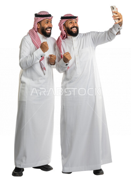 بورتريه لرجلان توأم عربيان خليجيان سعوديان، التقاط صورة تذكارية من خلال الهاتف المحمول، صور تذكارية، صور سيلفي، كاميرا امامية، مكالمة فيديو ، بث فيديو مباشر، ايماءات وجه ويدين تدل على الدهشة والحماس، صورة الجسم بالطول الكامل، خلفية بيضاء