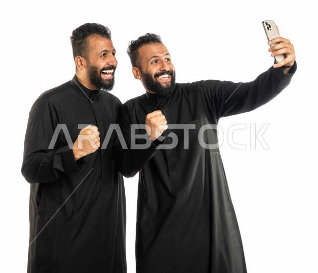بورتريه لرجلان توأم عربيان خليجيان سعوديان، التقاط صورة تذكارية من خلال الهاتف المحمول، صور تذكارية، صور سيلفي، كاميرا امامية، مكالمة فيديو، بث فيديو مباشر، ايماءات وجه ويدين تدل على الدهشة والحماس، خلفية بيضاء