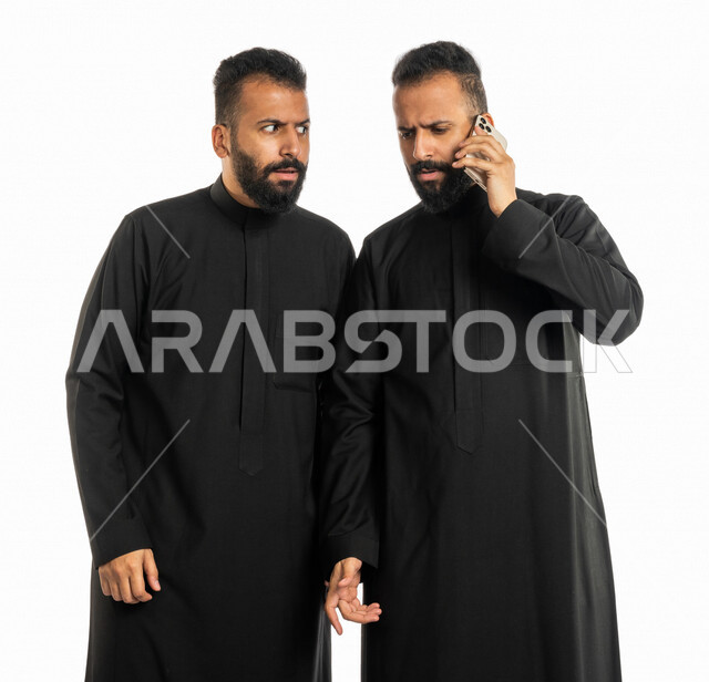 بورتريه لرجلان توأم عربيان خليجيان سعوديان، اجراء مكالمة من خلال الهاتف المحمول، الدهشة والاستغراب، مكالمة هاتفية، اخ ينظر الا اخوه نظرات تعجب، ايماءات وجه ويدين تدل على الدهشة والاستغراب، خلفية بيضاء