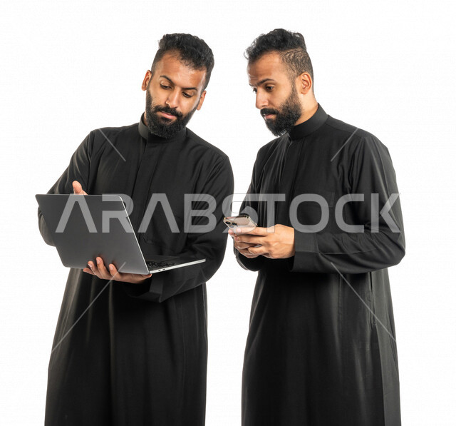بورتريه لرجلان توأم عربيان خليجيان سعوديان، اجتماع زملاء عمل في مقر عملهم، استخدام الحاسوب المحمول في تطوير العمل وحل المشكلات، استخدام الهاتف المحمول، توزيع مهام العمل، الوحده والعمل الجماعي، بيئة عمل سعودية، استخدام تقنيات حديثة، خلفية بيضاء