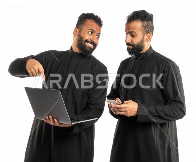 بورتريه لرجلان توأم عربيان خليجيان سعوديان، اجتماع زملاء عمل في مقر عملهم، استخدام الحاسوب المحمول في تطوير العمل وحل المشكلات، استخدام الهاتف المحمول، توزيع مهام العمل، الوحده والعمل الجماعي، بيئة عمل سعودية، استخدام تقنيات حديثة، خلفية بيضاء
