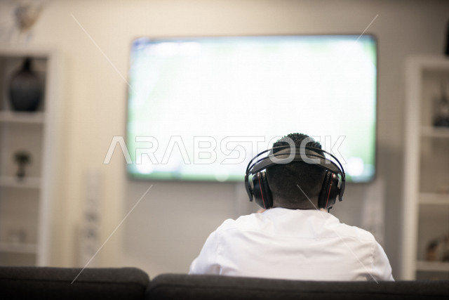 منظر من الخلف لشاب عربي سعودي خليجي يرتدي سماعة الرأس و يمسك بيده عصا التحكم و يلعب بألعاب الفيديو، المتعة و التسلية بألعاب الفيديو، ألعاب البلايستيشن الترفيهية، تقنيات و ألعاب, قضاء وقت ممتع في المنزل