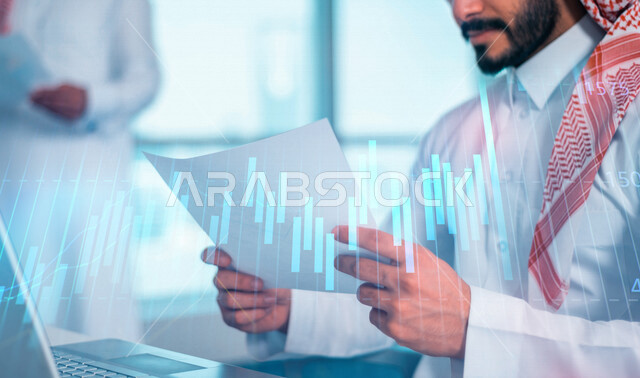 رجل عربي خليجي سعودي في مقر العمل، يحمل بيده ورقة لمخطط بياني مالي مدموجة بخلفية لرسومات بيانية لصعود وهبوط البورصة بتقنية الهولوجرام ثلاثية الأبعاد، الاستثمار المالي والاقتصادي، إستخدام جهازالحاسوب المحمول، دراسة السوق، مناقشة الرسم البياني المالي