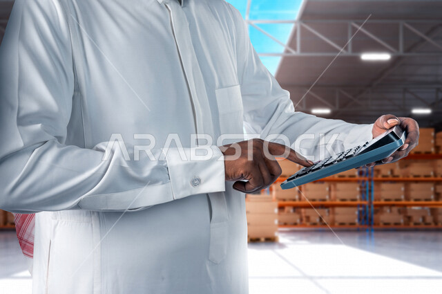 Close-up of a Saudi Arabian Gulf administrative supervisor, holding a calculator, working in the management of accounts inside the warehouse and stores, using the calculator in performing arithmetic operations, arithmetic professions and functions, the co