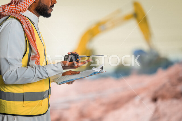 صورة مقربة لمهندس عربي خليجي سعودي، يرتدي الزي السعودي التقليدي وسترة الحماية خاصة بالعمل، يستخدم جهاز اتصال لاسلكي، يحمل بيده تقارير، متابعة العمل والإشراف على المشاريع، إعطاء أوامر خاصة بالعمل، مهن ووظائف هندسية، ادارة المشاريع الهندسية