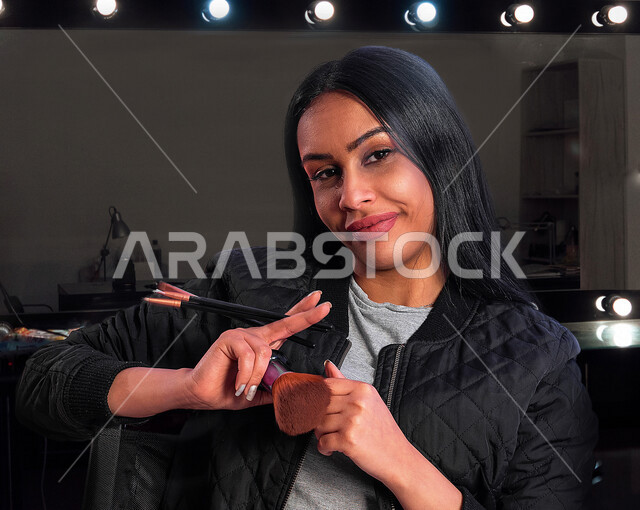 مرأة عربية خليجية سعودية تحمل مجموعة من فرش المكياج إحدى أدوات التجميل على بشرة الوجه، مرأة خبيرة بمستحضرات التجميل، التسويق لمنتجات التجميل، متجر بيع مستحضرات التجميل، مفهوم الإهتمام بالجمال والبشرة، ادوات مكياج تتوريال، صالون تجميلي نسائي