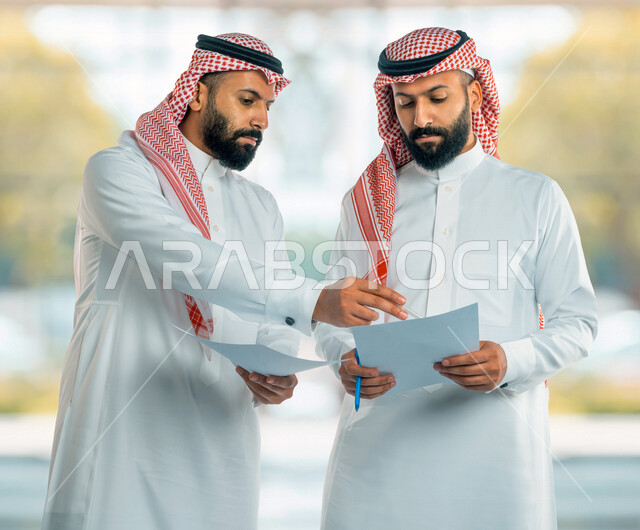 رجلان توأم عربيان خليجيان سعوديان يحملان اوراق عمل، مناقشة عمل، اجتماع زملاء العمل، تطوير العمل وحل المشكلات، توزيع مهام العمل، الوحده والعمل الجماعي، تطوير أهداف الشركة، اتفاقية عمل، بيئة عمل سعودية