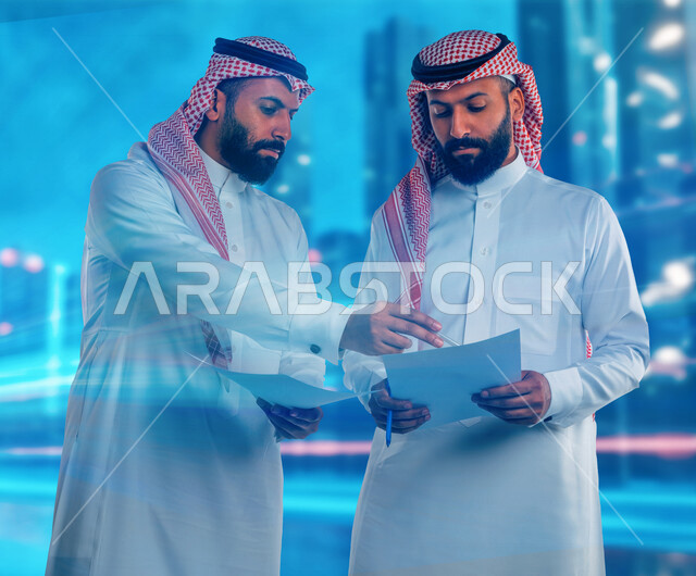 رجلان توأم عربيان خليجيان سعوديان يحملان اوراق عمل، مناقشة عمل، اجتماع زملاء العمل، تطوير العمل وحل المشكلات، توزيع مهام العمل، الوحده والعمل الجماعي، تطوير أهداف الشركة، اتفاقية عمل، بيئة عمل سعودية