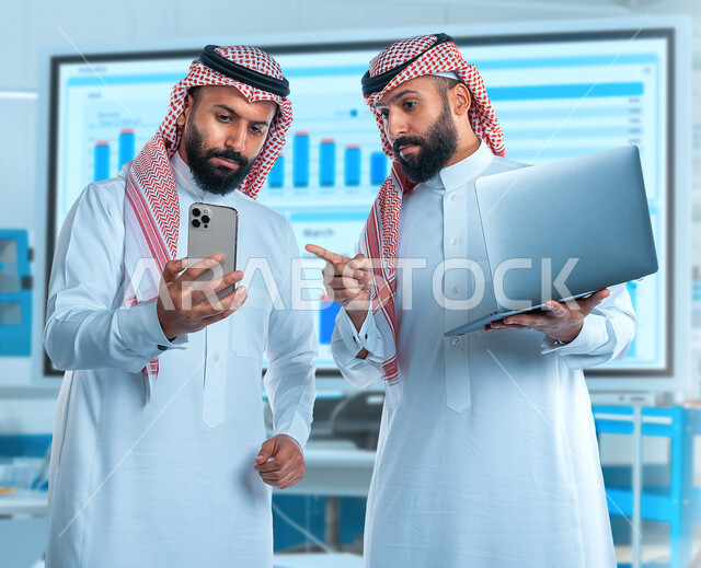 رجلان توأم عربيان خليجيان سعوديان، اجتماع زملاء عمل في مقر عملهم، استخدام جهاز الحاسوب والهاتف المحمول لمتابعة سوق الأوراق الماليه, التجاره الإلكترونيه, متابعة الأسهم, تطوير الشركات والمشاريع الإقتصاديه