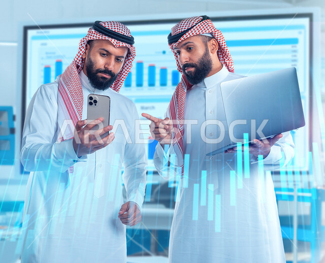 رجلان توأم عربيان خليجيان سعوديان، اجتماع زملاء عمل في مقر عملهم، استخدام جهاز الحاسوب والهاتف المحمول لمتابعة سوق الأوراق الماليه, التجاره الإلكترونيه, متابعة الأسهم, تطوير الشركات والمشاريع الإقتصاديه