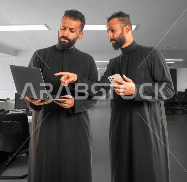 رجلان توأم عربيان خليجيان سعوديان، اجتماع زملاء عمل في مقر عملهم، استخدام جهاز الحاسوب والهاتف المحمول في تطوير العمل وحل المشكلات، توزيع مهام العمل، الوحده والعمل الجماعي، بيئة عمل سعودية، استخدام تقنيات حديثة
