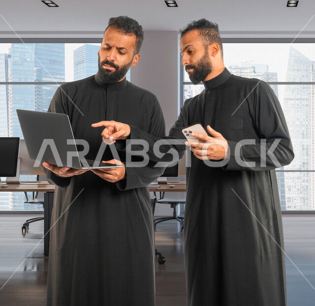 رجلان توأم عربيان خليجيان سعوديان، اجتماع زملاء عمل في مقر عملهم، استخدام جهاز الحاسوب والهاتف المحمول في تطوير العمل وحل المشكلات، توزيع مهام العمل، الوحده والعمل الجماعي، بيئة عمل سعودية، استخدام تقنيات حديثة