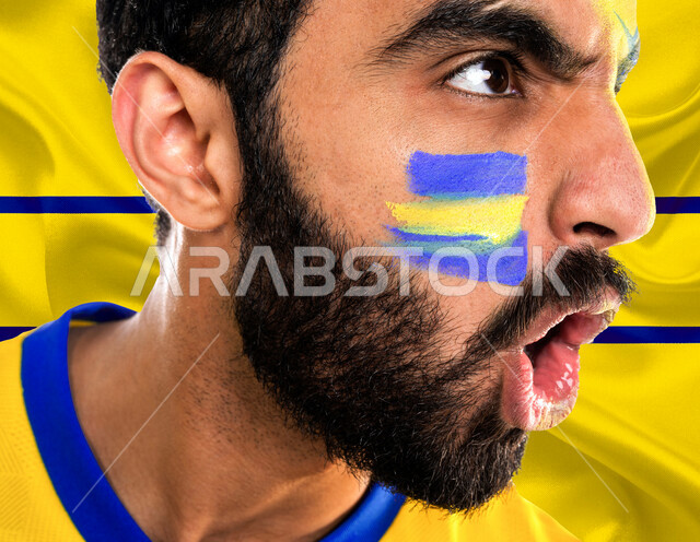 شاب عربي خليجي سعودي يرتدي تيشرت نادي النصر السعودي، تشجيع فريق النصر السعودي لكرة قدم، متابعة مباراة كرة قدم، شاب يرسم على وجهه شعار نادي النصر السعودي، إيماءات وجه ويدين تدل على السعادة والحماس والشغف بالفوز