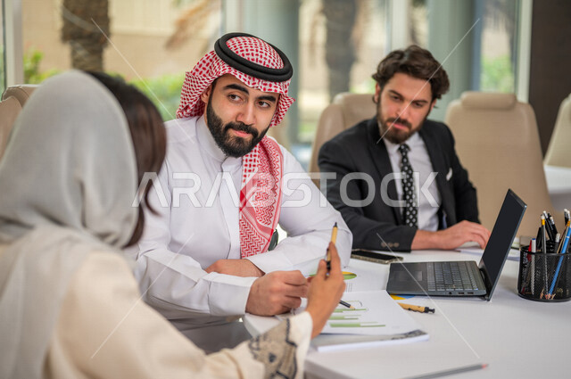A Saudi Arabian Gulf businessman in the traditional Saudi dress in a business meeting with a new client wearing a formal suit, a Saudi Arabian Gulf business woman working on the study and analysis of new projects, the use of a laptop in business developme