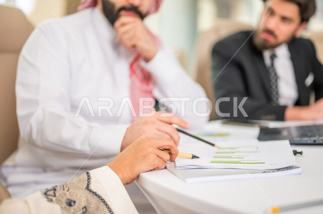 رجل اعمال عربي سعودي خليجي بالثوب السعودي التقليدي في اجتماع عمل مع عميل جديد يرتدي البدلة الرسمية، سيدة اعمال عربية سعودية خليجية تعمل على دراسة وتحليل المشاريع الجديدة، استخدام الحاسوب المحمول في تطوير العمل، اجتماعات عمل، الشراكة والتعاون