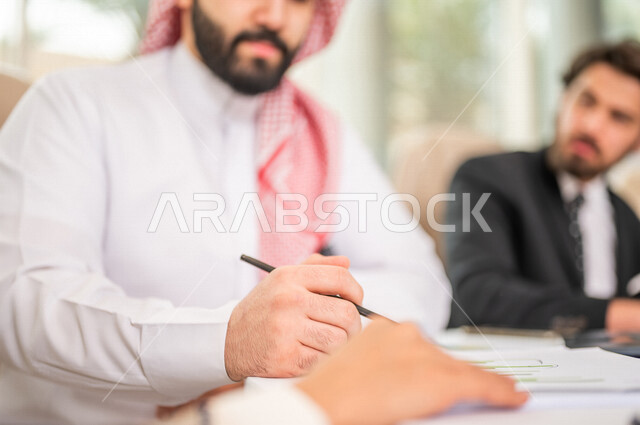 A Saudi Arabian Gulf businessman in the traditional Saudi dress in a business meeting with a new client wearing a formal suit, using a laptop computer in work development and problem solving, business meetings, project development, study and analysis of n