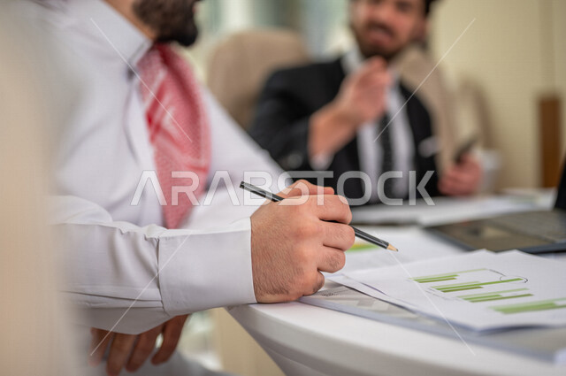رجل اعمال عربي سعودي خليجي بالثوب السعودي التقليدي في اجتماع عمل مع عميل جديد يرتدي البدلة الرسمية، استخدام الحاسوب المحمول في تطوير العمل وحل المشكلات، اجتماعات عمل، تطوير المشاريع، دراسة وتحليل المشاريع الجديدة، الشراكة والتعاون 
