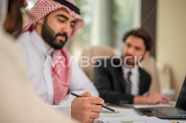 رجل اعمال عربي سعودي خليجي بالثوب السعودي التقليدي في اجتماع عمل مع عميل جديد يرتدي البدلة الرسمية، سيدة اعمال عربية سعودية خليجية تعمل على دراسة وتحليل المشاريع الجديدة، استخدام الحاسوب المحمول في تطوير العمل، اجتماعات عمل، الشراكة والتعاون