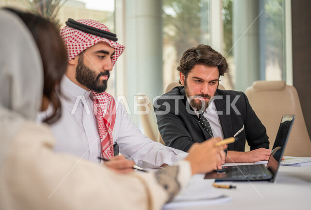 رجل اعمال عربي سعودي خليجي بالثوب السعودي التقليدي في اجتماع عمل مع عميل جديد يرتدي البدلة الرسمية، سيدة اعمال عربية سعودية خليجية تعمل على دراسة وتحليل المشاريع الجديدة، استخدام الحاسوب المحمول في تطوير العمل، اجتماعات عمل، الشراكة والتعاون