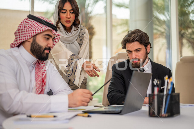 رجل اعمال عربي سعودي خليجي بالثوب السعودي التقليدي في اجتماع عمل مع عميل جديد يرتدي البدلة الرسمية، سيدة اعمال عربية سعودية خليجية تعمل على دراسة وتحليل المشاريع الجديدة، استخدام الحاسوب المحمول في تطوير العمل، اجتماعات عمل، الشراكة والتعاون