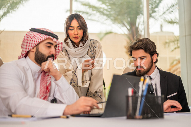 رجل اعمال عربي سعودي خليجي بالثوب السعودي التقليدي في اجتماع عمل مع عميل جديد يرتدي البدلة الرسمية، سيدة اعمال عربية سعودية خليجية تعمل على دراسة وتحليل المشاريع الجديدة، استخدام الحاسوب المحمول في تطوير العمل، اجتماعات عمل، الشراكة والتعاون